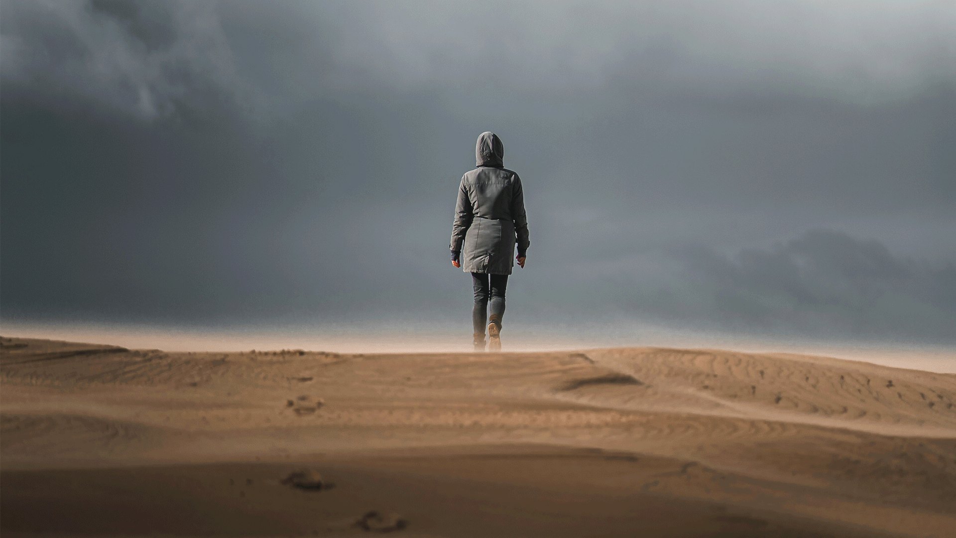 Lone hooded figure standing on wind-swept sand under a stormy sky, before masking in Lightroom