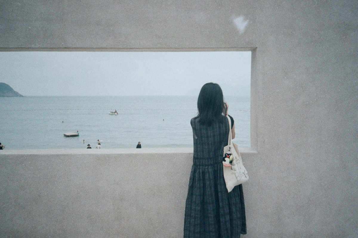 A woman in a plaid dress looking out a window opening at the sea, with people and boats in the water before using the Remove tool