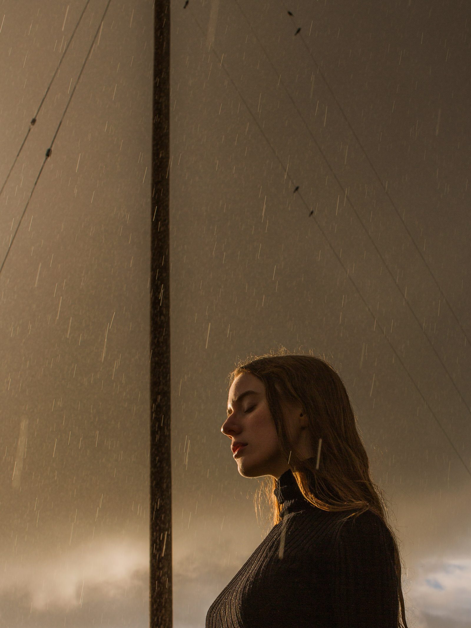 Woman in a black sweater with her eyes closed under rain and warm light, a utility pole and wires against a gray sky