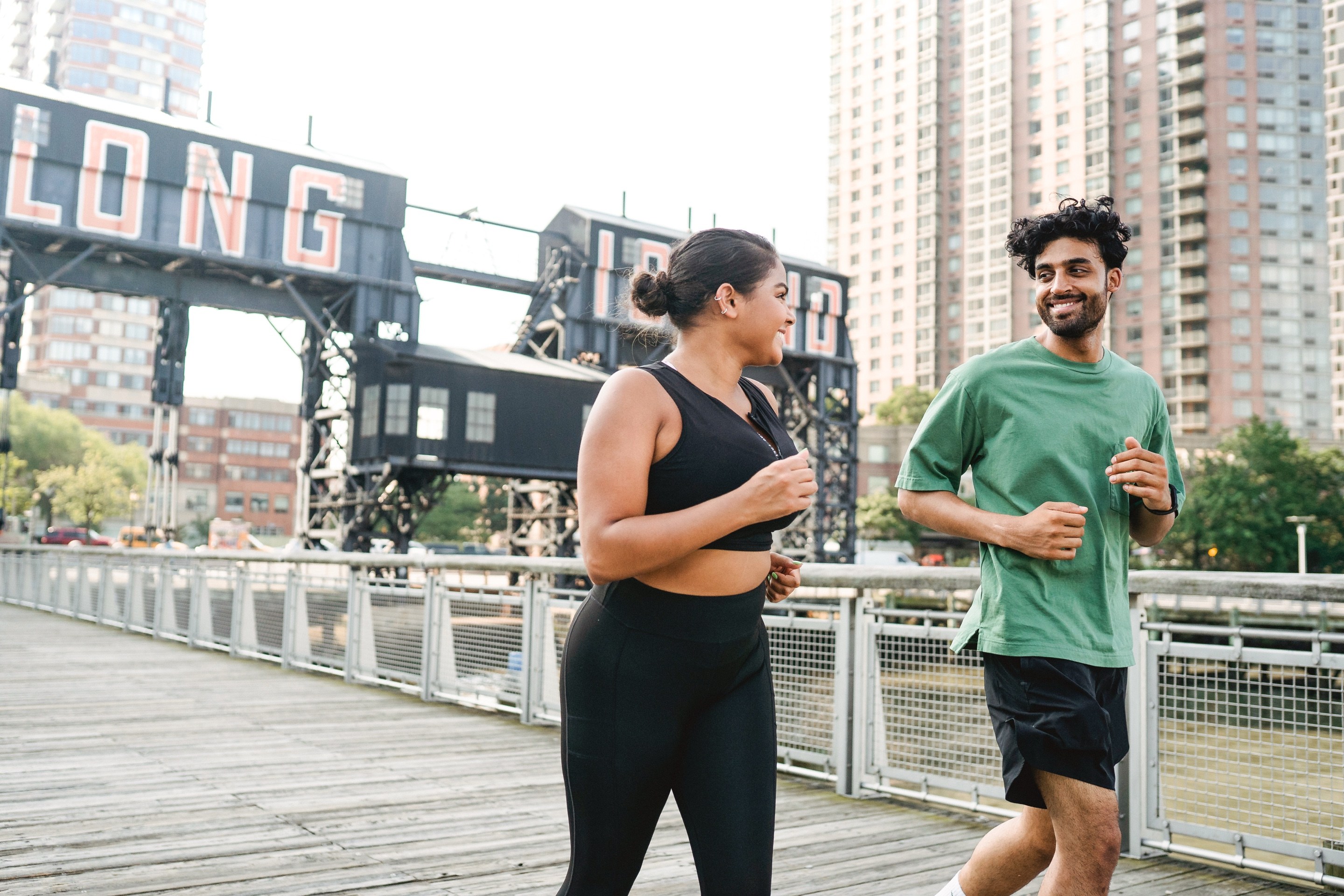 Two people jogging together outdoors, smiling and talking, near a historic industrial gantry structure with city high-rises behind them, from a tutorial on the fastest way to edit multiple photos in Lightroom Classic.
