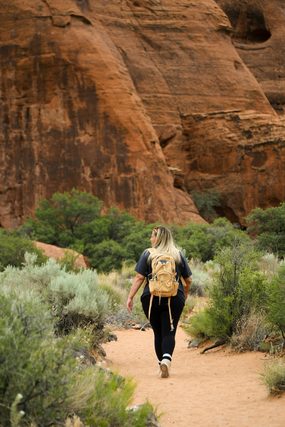 Woman with a backpack walking a sandy trail below red cliffs