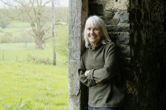 Smiling older woman leaning in a stone doorway by a field