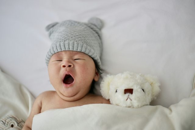 Yawning baby in a grey knit hat beside a teddy bear