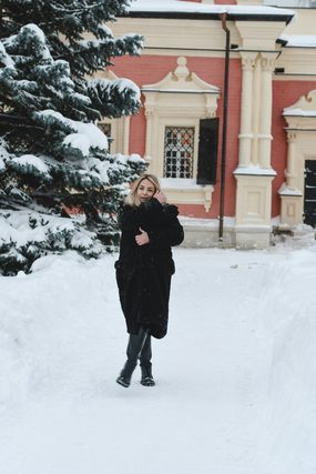 Woman in a black fur coat on a snowy path by a red building
