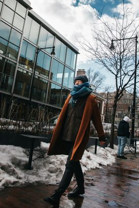 Man in an orange coat and blue scarf walking past a glass building in winter