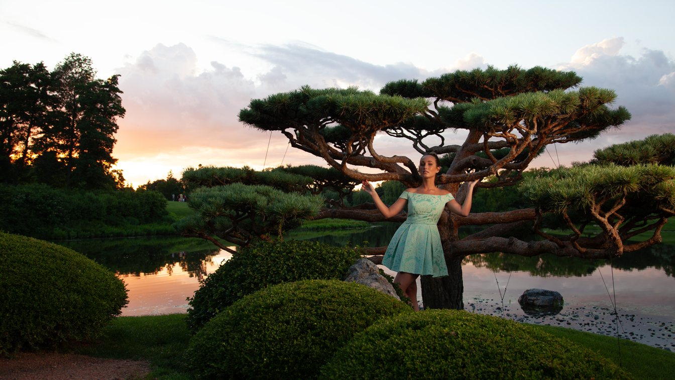 A woman in a mint dress under a sculpted pine tree beside a pond at sunset, with a warm cast over her skin and a pale sky
