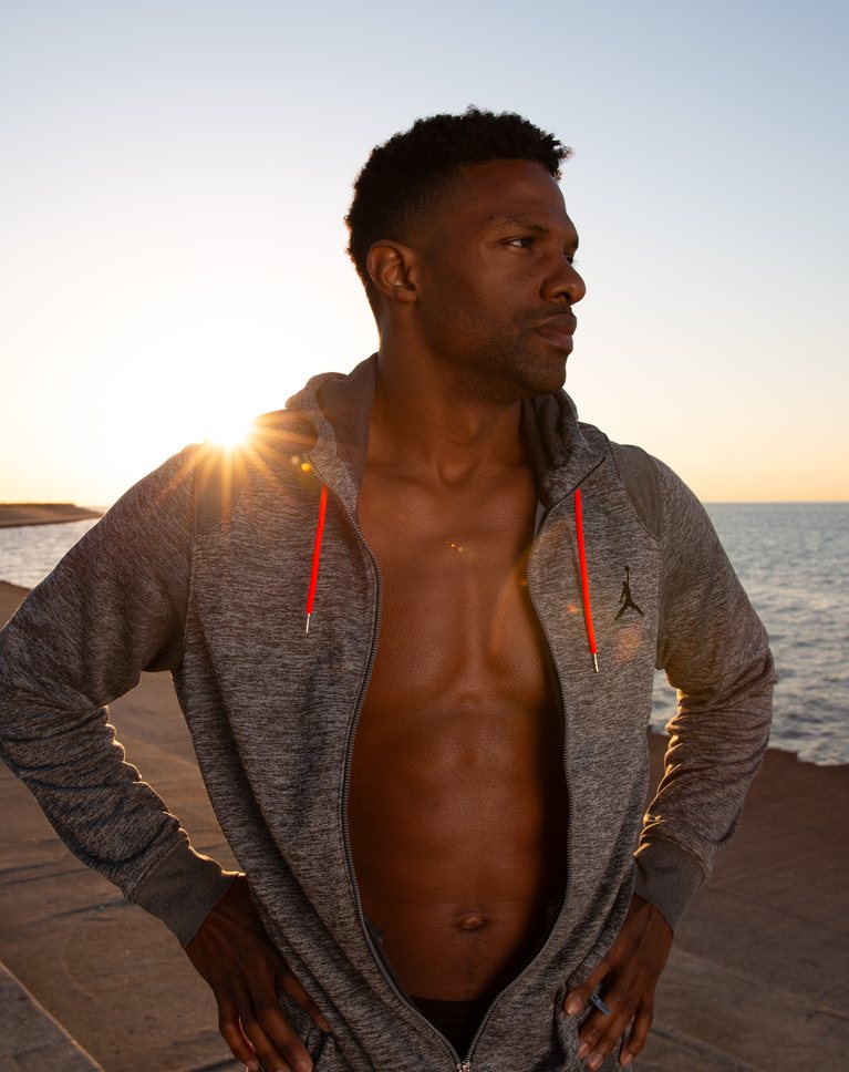 A man in a gray hoodie on a lakefront at sunrise, with flat skin and a hazy sky