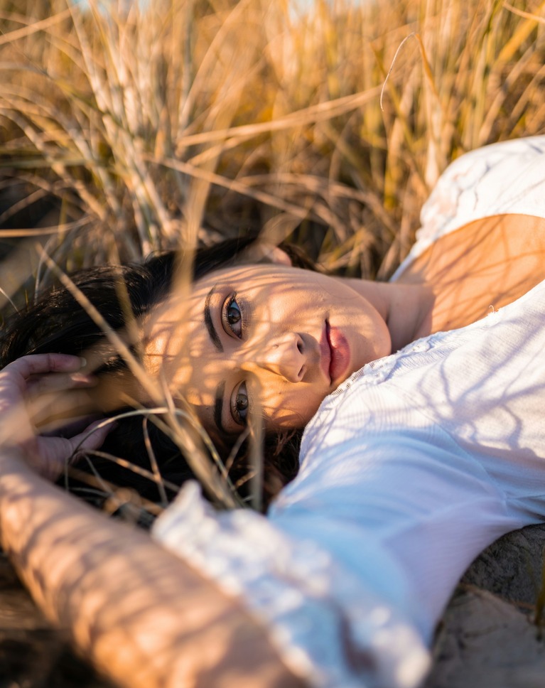 A woman lying in dry beach grass in late sun, with grass shadows across her face and a yellow-green cast on her skin, before correction