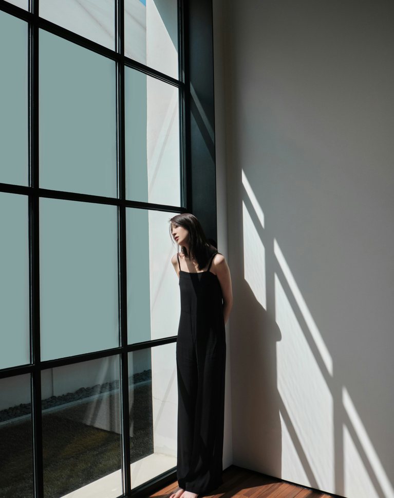 A woman in black standing beside a tall window wall in cool, neutral light
