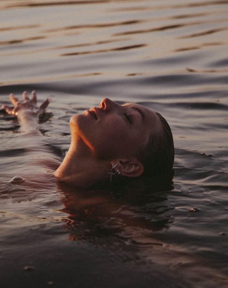 A woman floating on her back in calm water at sunset, eyes closed, before the color grade