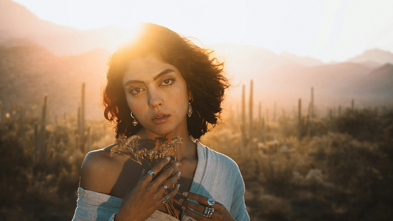 A woman with short curly hair holding dried flowers in the desert at sunset, with cacti and mountains behind her, before the color grade