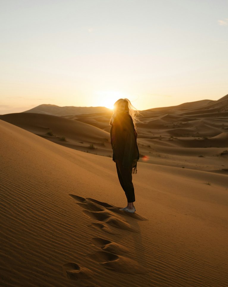 A woman walking on desert dunes toward a low sun under a pale, hazy sky