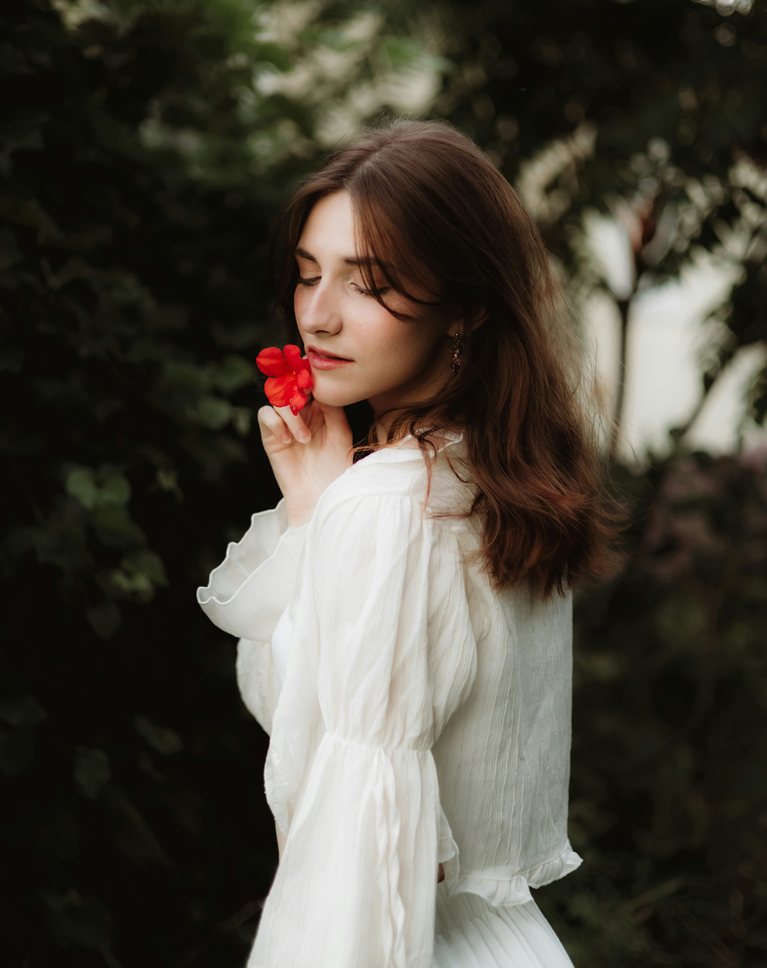 A woman in a white blouse holding a red flower in front of dark green leaves