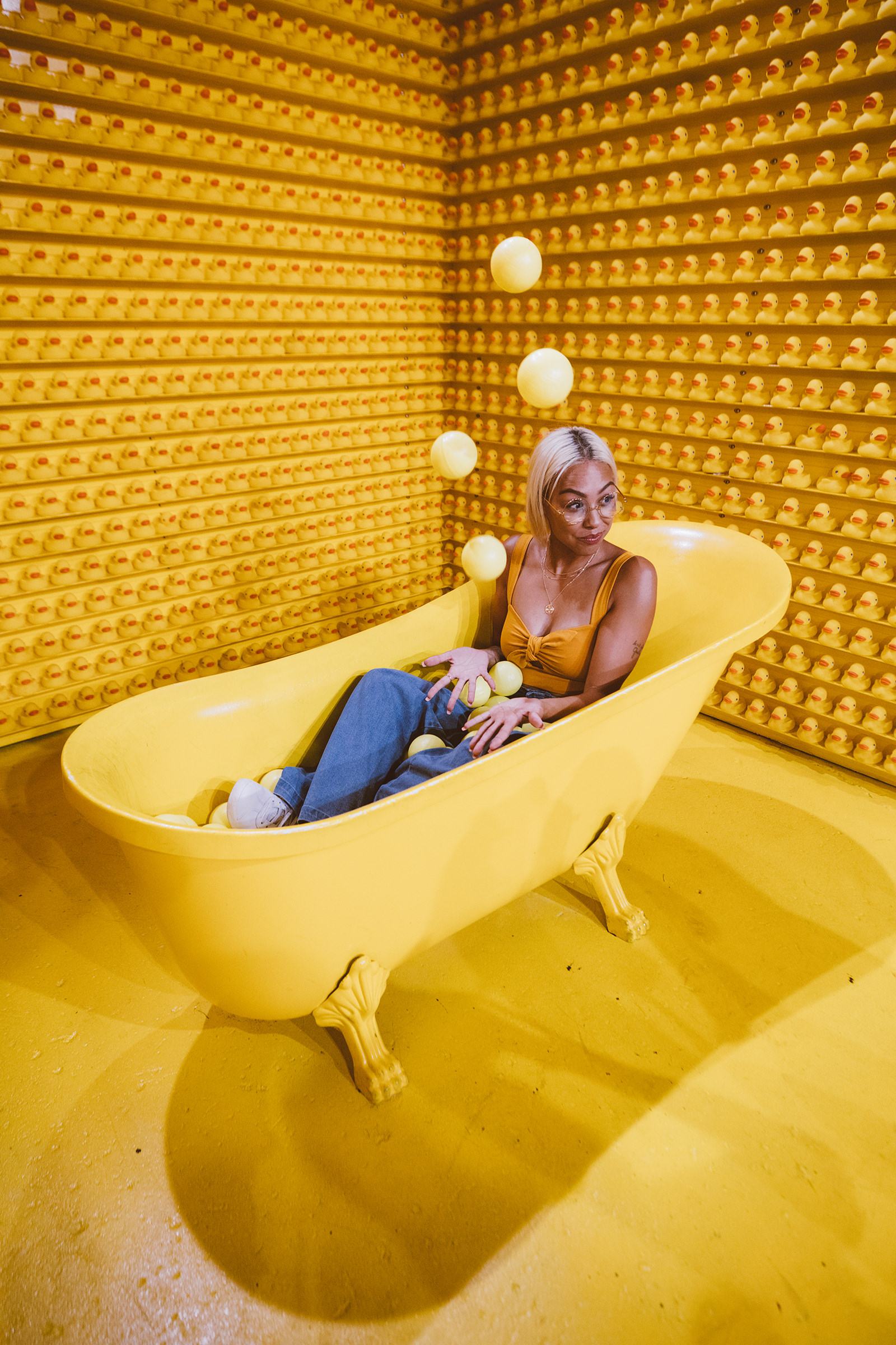 Woman in a yellow top sits in a yellow clawfoot tub in a room lined with rubber ducks, yellow balls floating around her