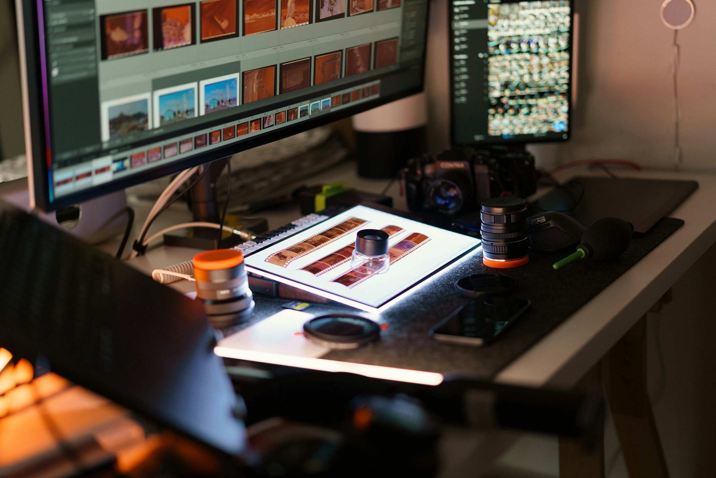 Affinity vs Photoshop: a photographer's desk with two monitors of photo thumbnails, a film camera, lenses and a light table with strips of film