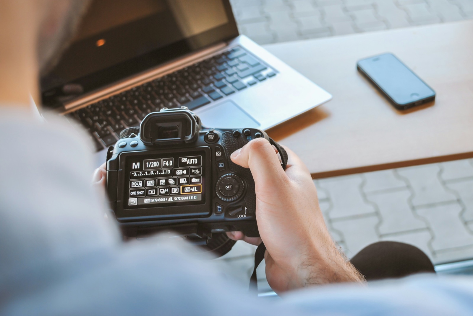 A photographer holding a Canon DSLR with its settings screen showing manual mode, 1/200 second and f/4