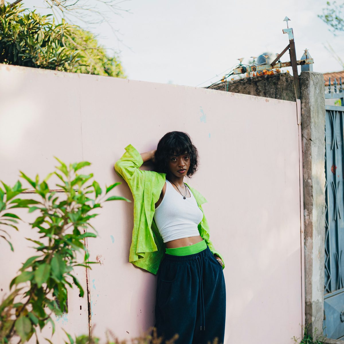 A woman in a lime green jacket leaning against a pale pink wall with green plants in front