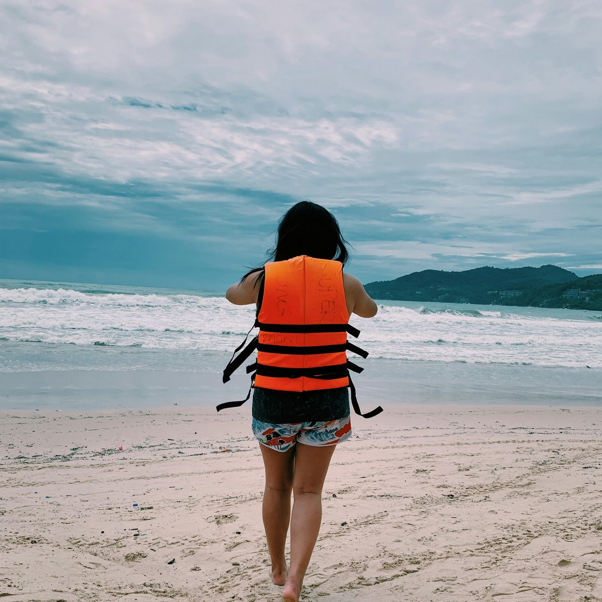 A woman in an orange life jacket seen from behind walking toward a grey-blue sea under an overcast sky