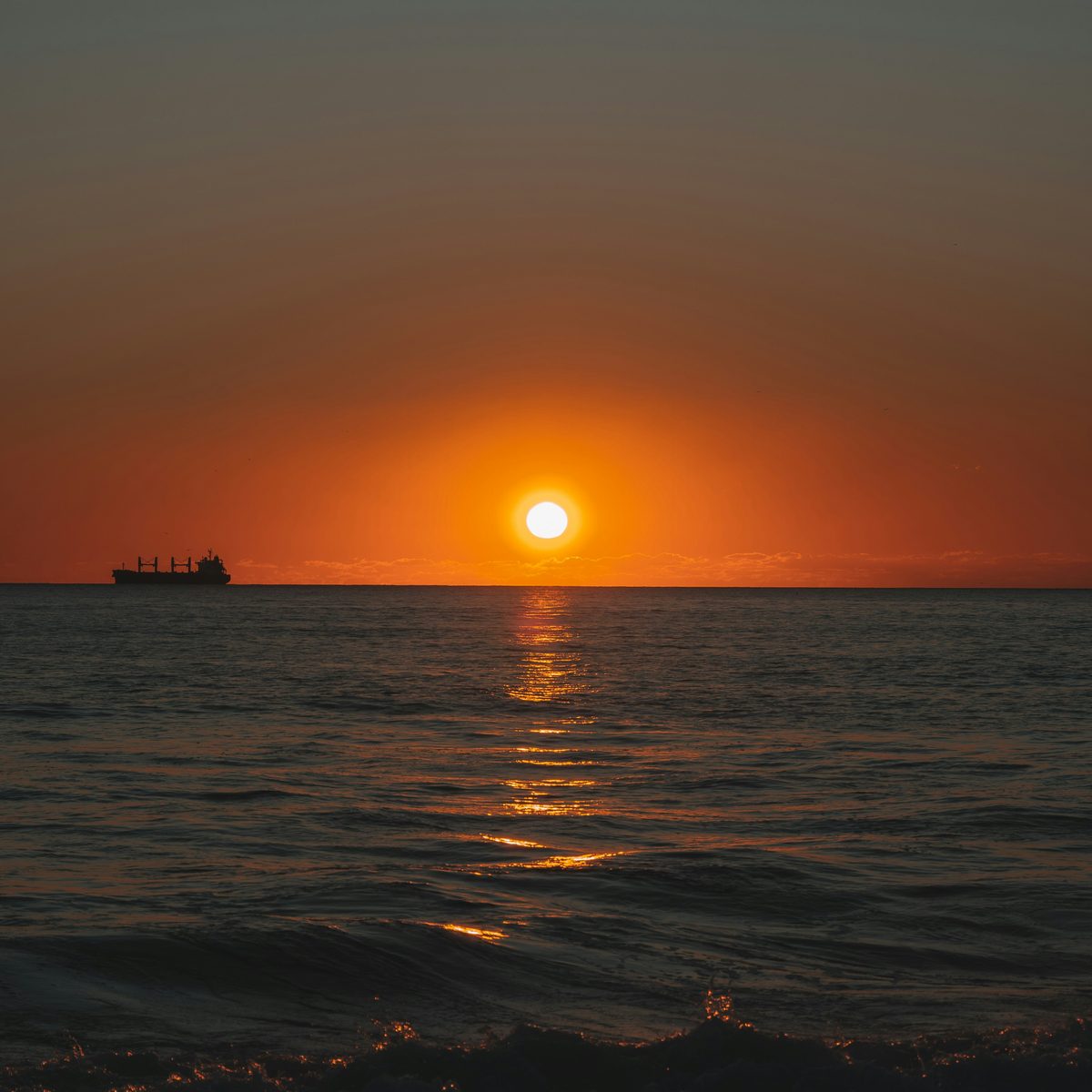 The sun setting into a dark blue-grey sea under an orange sky, with a ship on the horizon