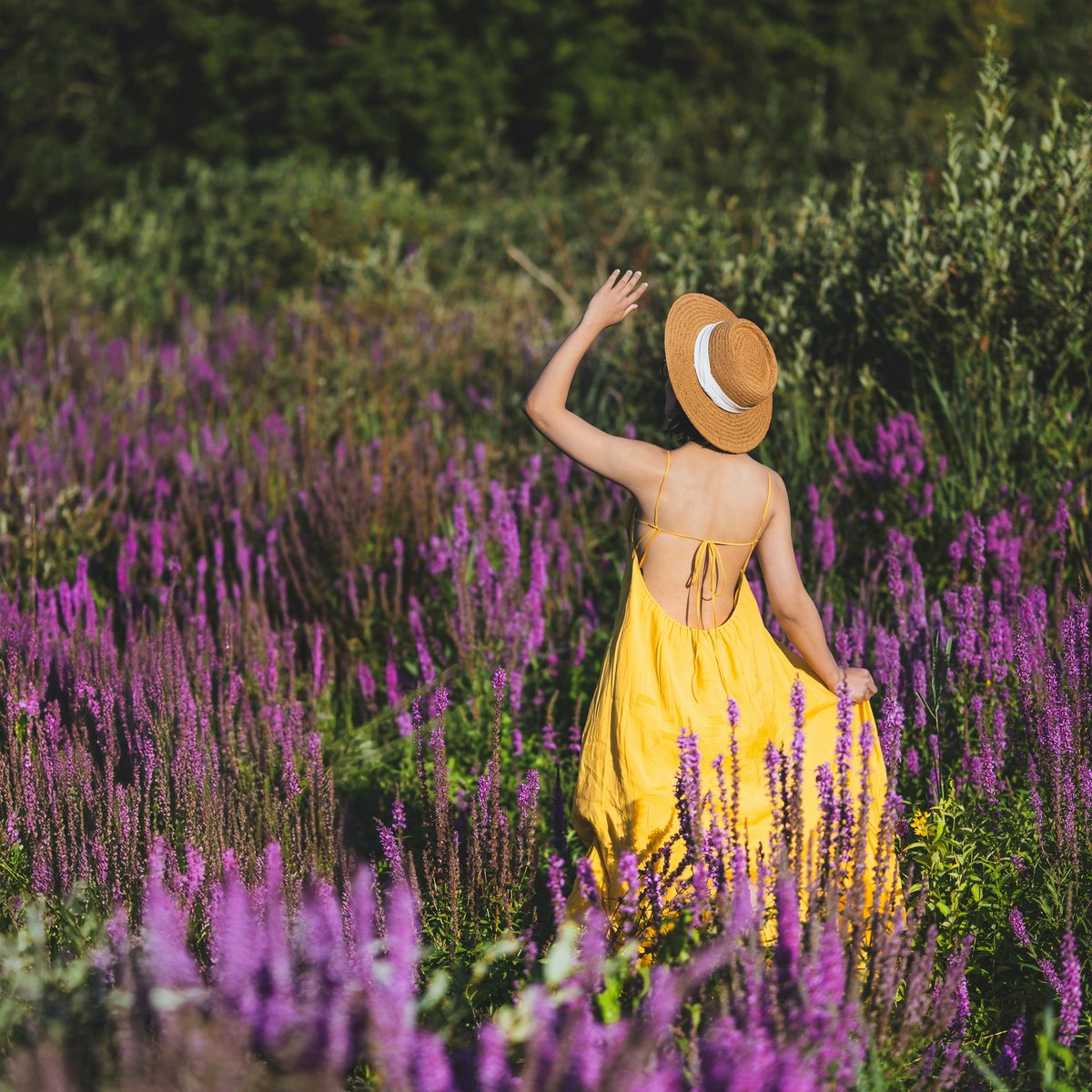 A woman in a yellow dress and tan straw hat standing with one arm raised in a field of purple flowers