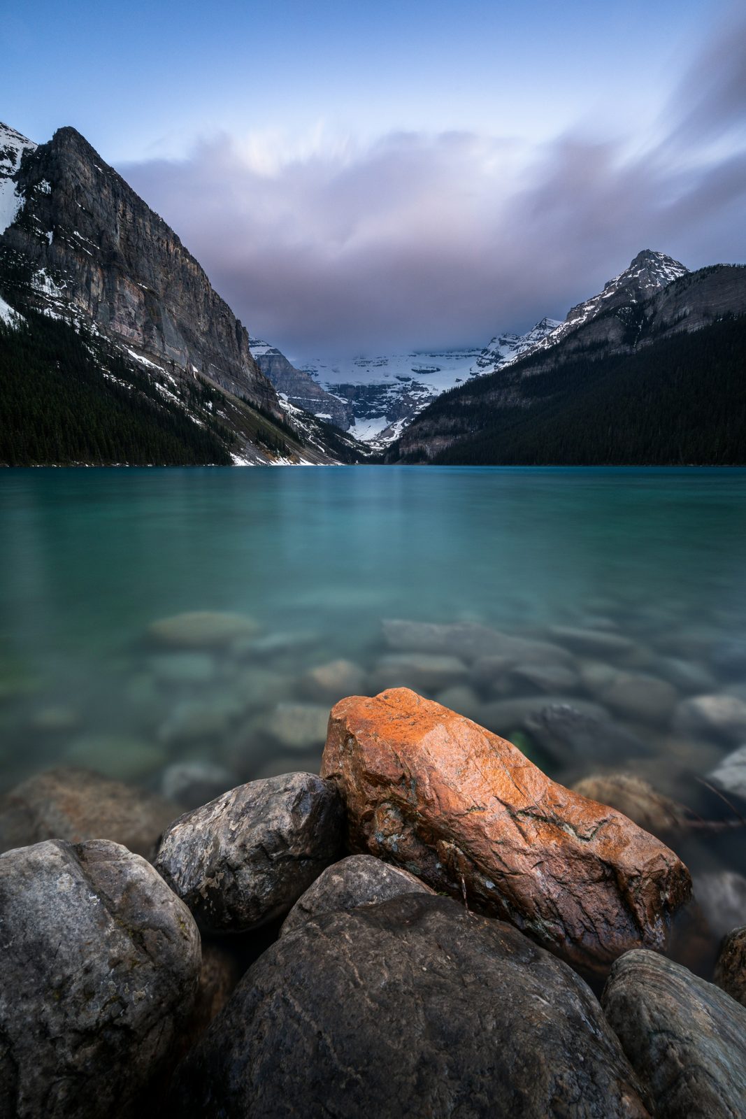 An orange rock in the foreground, a turquoise lake behind it and mountains in the distance