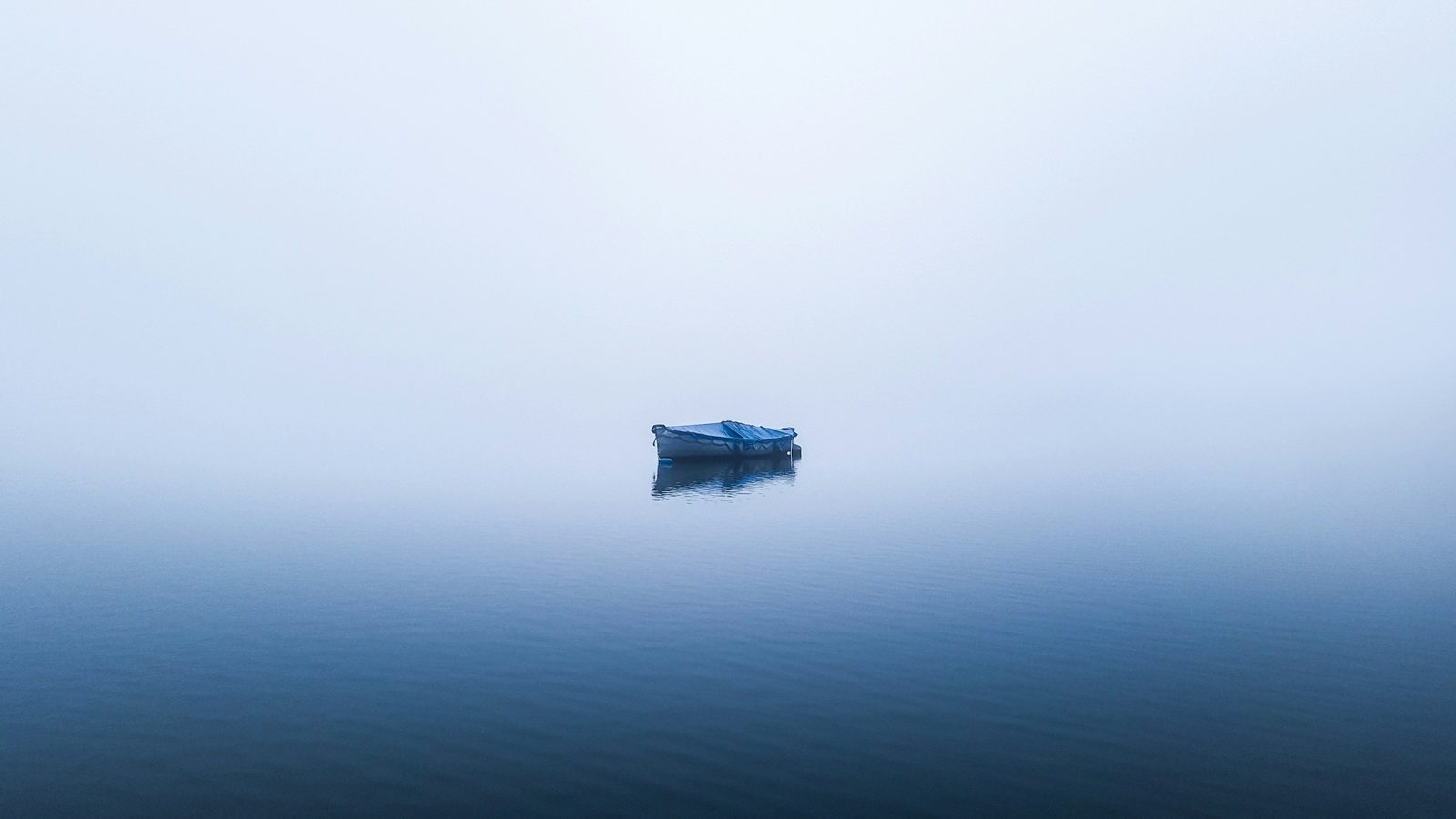 A lone boat under a blue cover on a vast expanse of pale water and fog