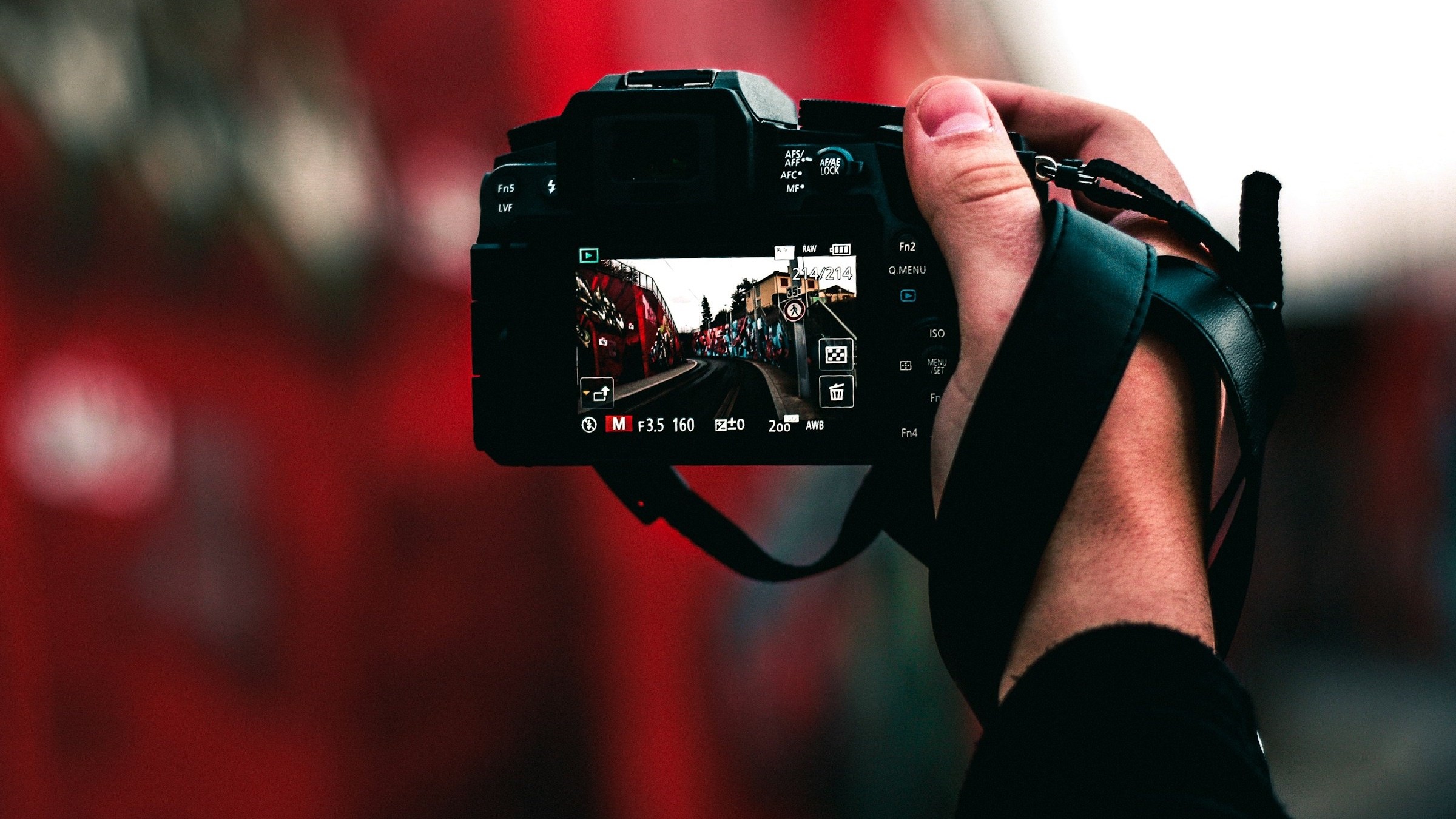 The back of a camera showing a street photo on its screen, held up against a red wall