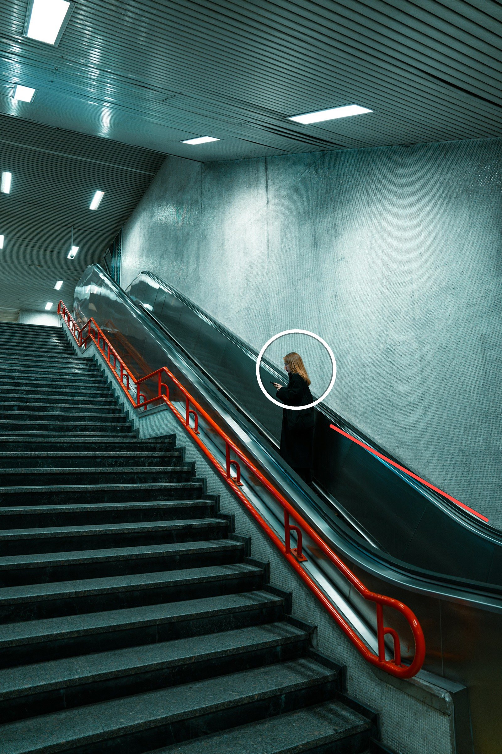 A woman riding an escalator in an underground station, with the escalator rail and a red handrail running diagonally toward her