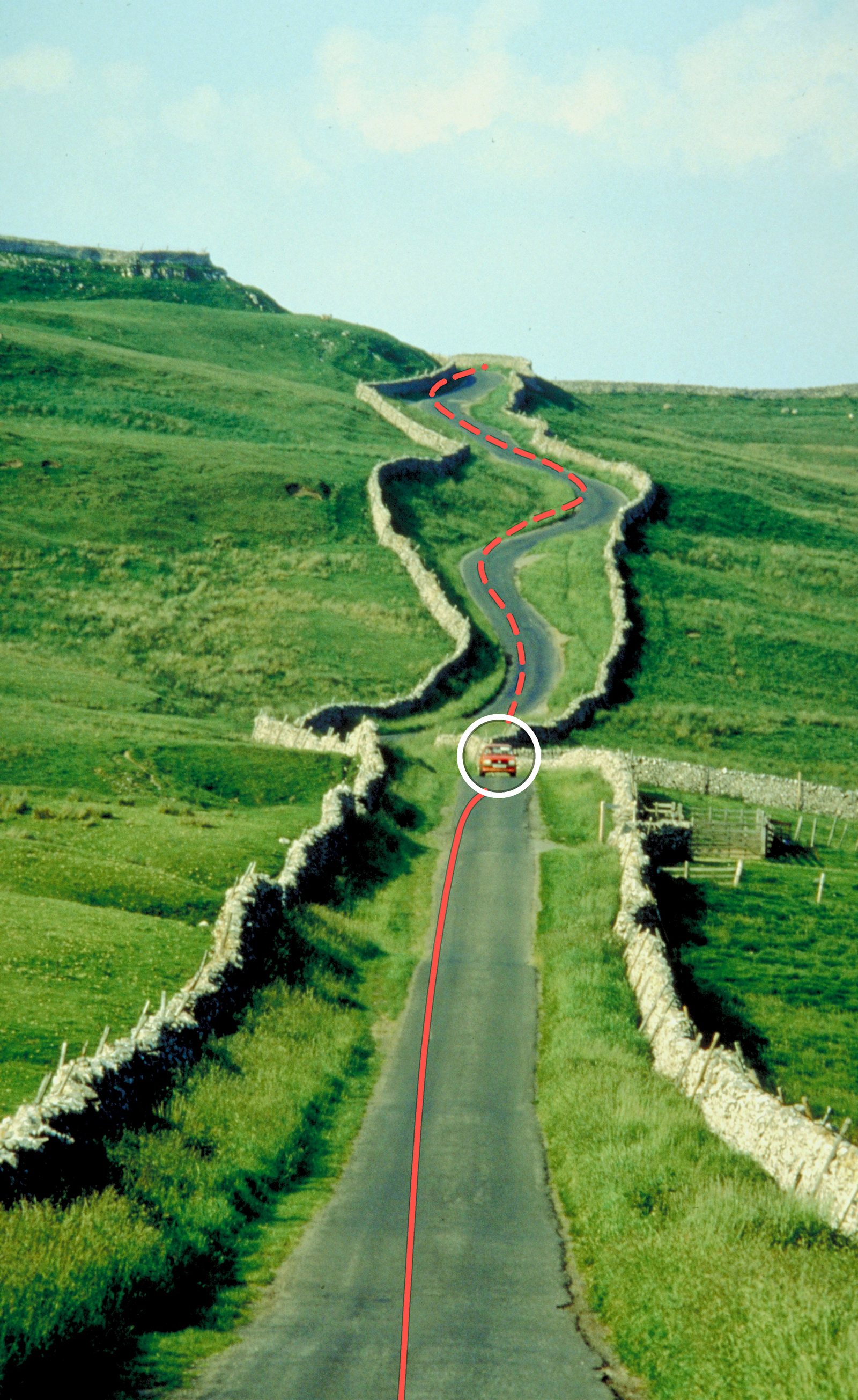 A narrow road winding between stone walls up a green hillside, with one red car on it
