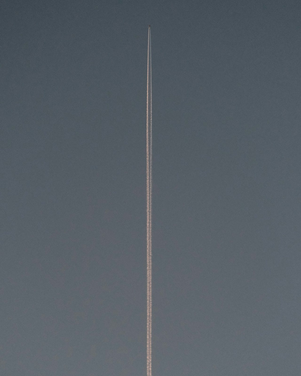 A contrail drawn straight up a plain grey sky