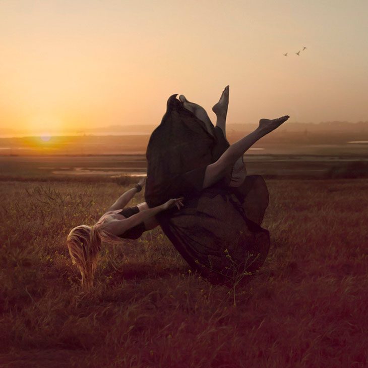 A blonde woman in a long dark dress floats upside down above a grassy field at sunset, legs in the air