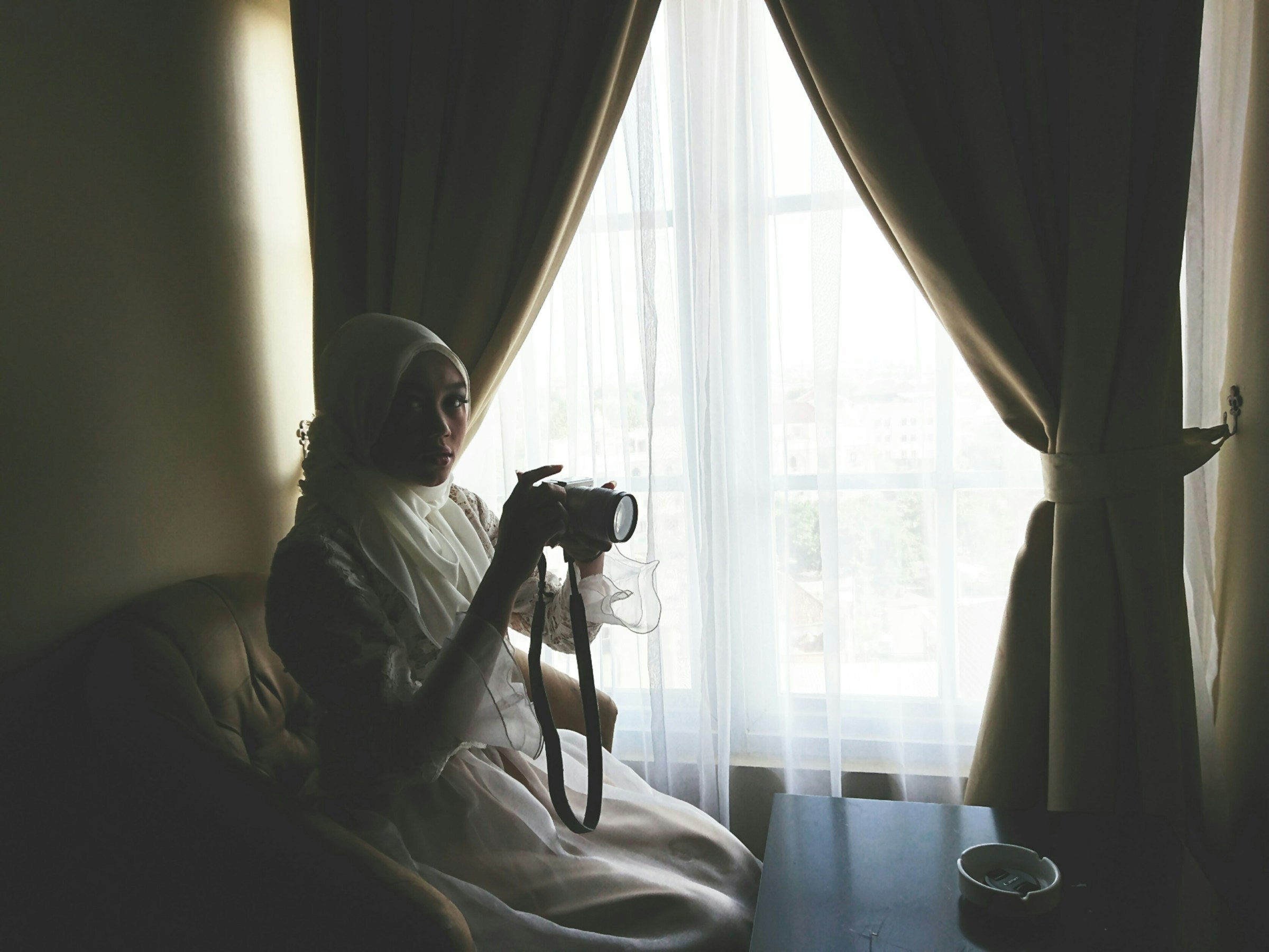 Photographer sitting by a curtained window at home, holding a camera