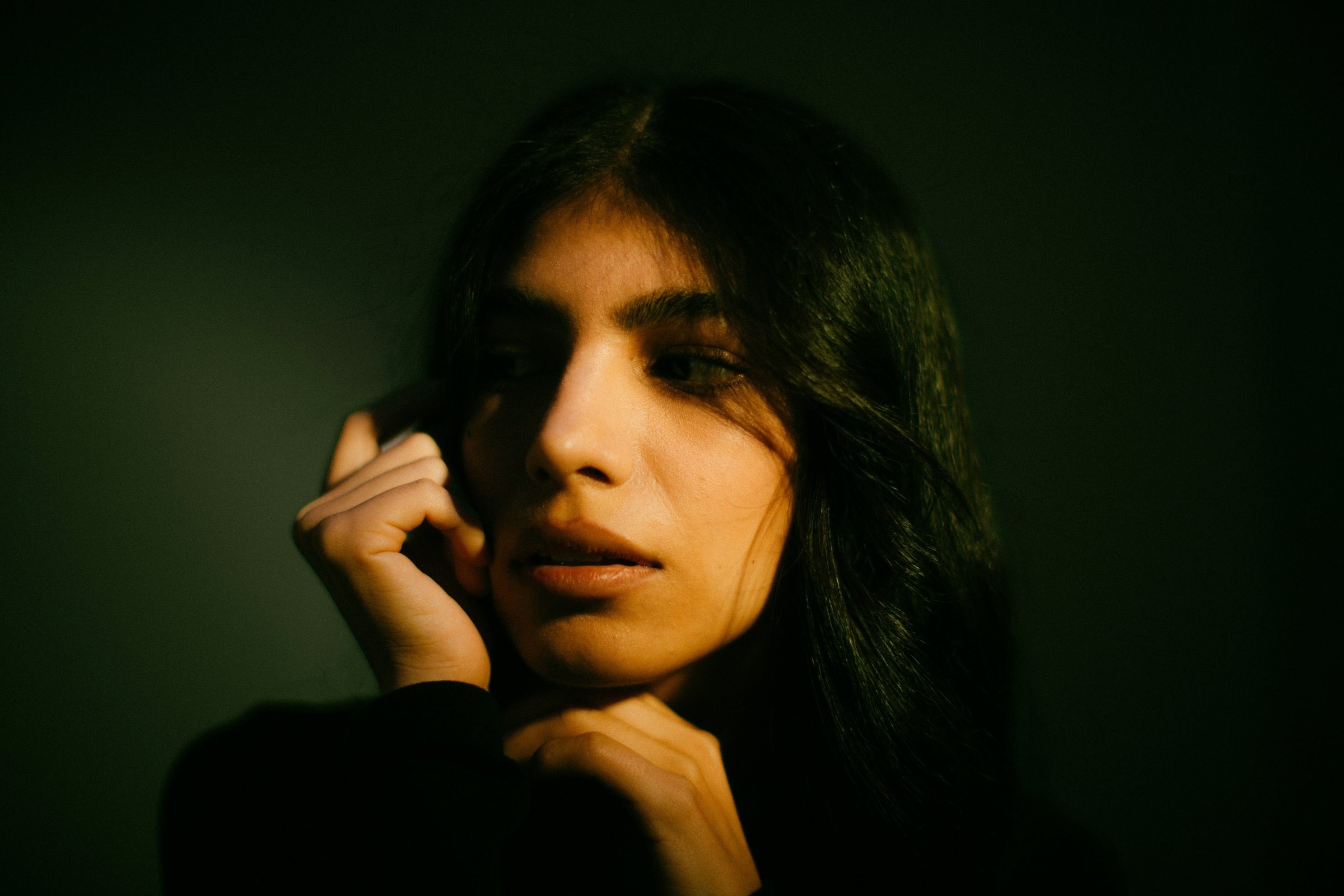 Portrait lit by a single warm light from one side, with half of her face falling into shadow against a dark green wall